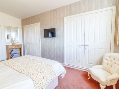 A bedroom with a bed a patterned bed runner a white armchair a wooden cabinet with towels a wall mirror and a mounted television at The Croft in Leigh Sinton