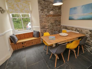 A dining area with a wooden table yellow and gray chairs stone wall and wooden bench with cushions at Fairfield Cottage in Boscastle