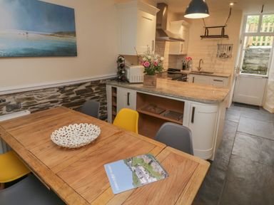 A kitchen with a wooden dining table yellow and grey chairs and a countertop with flowers at Fairfield Cottage in Boscastle