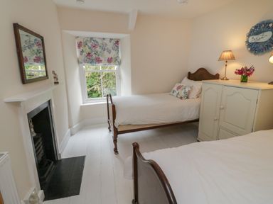 A bedroom with two single beds a fireplace a window with floral blinds and a cabinet with lamps and flowers at Fairfield Cottage in Boscastle