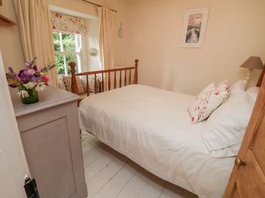 A bedroom with a wooden bed and white bedding a dresser with flowers and a window seat with cushions at Fairfield Cottage in Boscastle