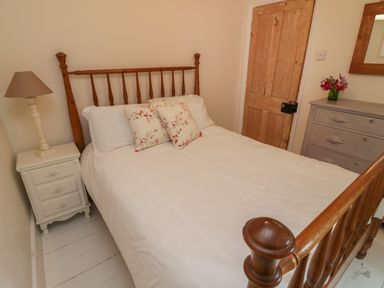A bedroom with a wooden bed frame white bedding floral pillows a nightstand with a lamp wooden door a dresser with a mirror and flowers at Fairfield Cottage in Boscastle