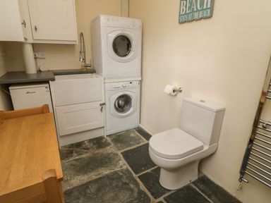 A utility room with a stacked washer and dryer a sink a toilet a wooden table a towel radiator and a sign on the wall at Fairfield Cottage in Boscastle