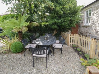 A garden patio with a metal table and six chairs surrounded by plants and a wooden fence at Fairfield Cottage in Boscastle