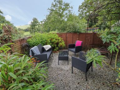 An outdoor seating area with black wicker furniture on a gravel ground surrounded by plants at Fairfield Cottage in Boscastle