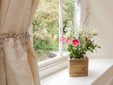 A bouquet of flowers in a wooden vase on a windowsill next to a cream curtain held with a beaded tieback at Fairfield Cottage in Boscastle