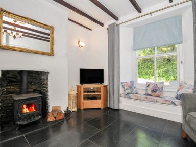 A living room with a wood burning stove a television on a wooden stand and a window seat with cushions at Fairfield Cottage in Boscastle