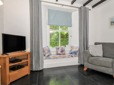 A living room with a gray sofa gray curtains a wooden TV stand with a television and a window bench with cushions at Fairfield Cottage in Boscastle