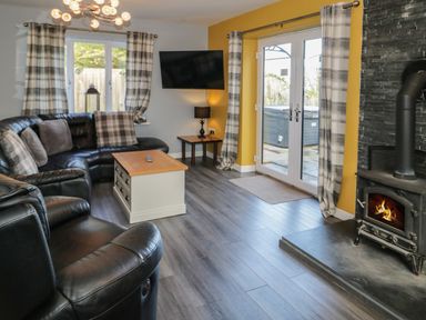 A living room with black leather sofas a wooden coffee table a wall-mounted television and a wood-burning stove at Big Azzles in Tyn-y-Gongl near Benllech