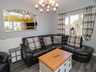 A living room with a black leather corner sofa with cushions a wooden coffee table a large decorative mirror wall radiator and window with plaid curtains at Big Azzles in Tyn-y-Gongl near Benllech