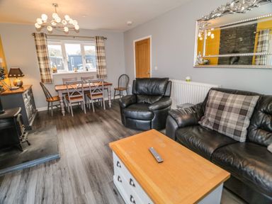 A living room with black leather sofa and armchair a wooden coffee table and a dining table with chairs by a window at Big Azzles in Tyn-y-Gongl near Benllech