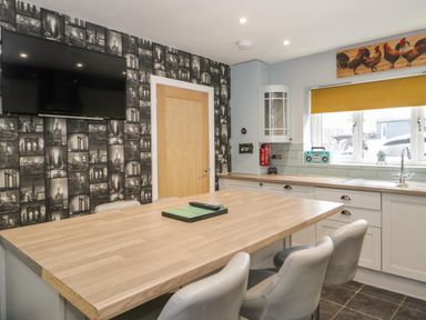A kitchen with a wooden island and chairs a wall-mounted TV and a window with a yellow blind at Big Azzles in Tyn-y-Gongl near Benllech