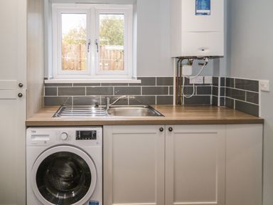 A utility room with a washing machine under a wooden countertop with a sink and a boiler on the wall at Big Azzles in Tyn-y-Gongl near Benllech
