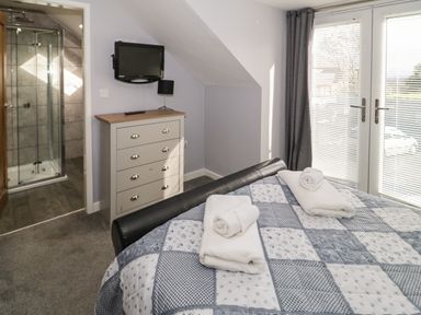 A bedroom with a bed covered with a patterned quilt and towels on top a chest of drawers with a lamp and a wall-mounted television a glass shower enclosure is visible through an open door and double doors with blinds lead outside at Big Azzles in Tyn-y-Gongl near Benllech