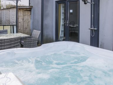 An outdoor patio area with a bubbling hot tub a wicker dining set and an outdoor shower near a gray building at Big Azzles Tyn-y-Gongl near Benllech