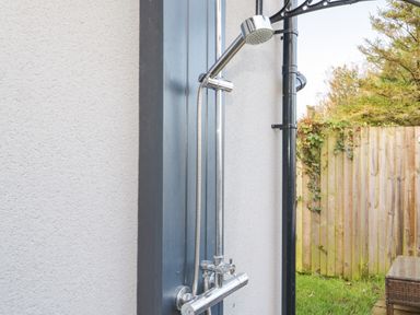 An outdoor shower installed on an exterior wall next to a garden with a wooden fence at Big Azzles in Tyn-y-Gongl near Benllech