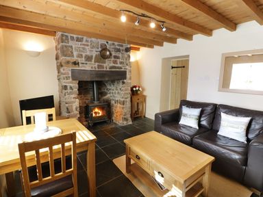 A living room with a stone fireplace wooden coffee table black leather sofa and wooden dining table at Menai Cottage in Brynsiencyn