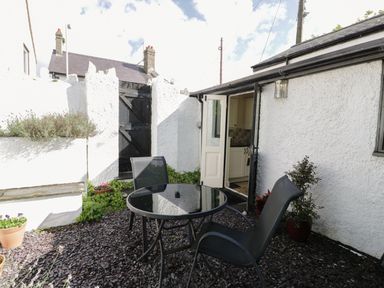A small outdoor patio with two black chairs and a round glass table near the open door of a white building at Menai Cottage in Brynsiencyn
