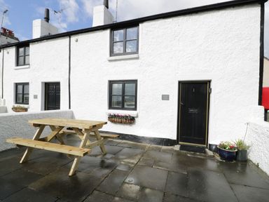 Exterior view of a white stone cottage with black doors and windows a wooden picnic table on the stone patio at Menai Cottage in Brynsiencyn