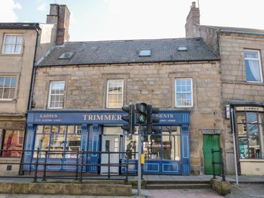 A street view of a stone building with a blue shopfront for a trimmers barbershop and a pedestrian crossing signal at 1 Pringles Yard in Alnwick