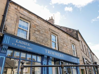 A stone building with blue storefront signage for ladies and gents trimmers at 3-5 Clayport Street at 1 Pringles Yard in Alnwick