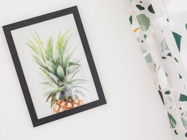 A framed picture of a pineapple on a white wall next to a curtain with a colorful abstract pattern at 1 Pringles Yard in Alnwick