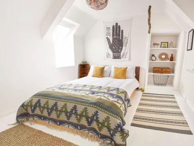 A bedroom with a bed covered by a patterned blanket two yellow pillows a wall hanging a bookshelf and rugs on the floor at 1 Pringles Yard in Alnwick