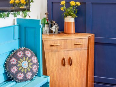 A room corner with a small wooden cabinet with bottles and a vase of yellow flowers next to a blue bench with a decorative round cushion at 1 Pringles Yard in Alnwick