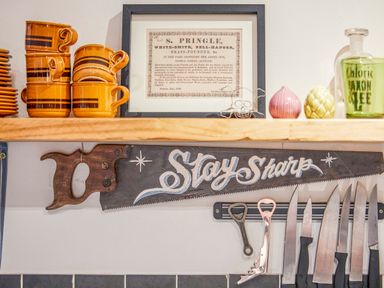 A wooden shelf with stacked orange cups plates and framed paper above a saw with stay sharp written and knives on a magnetic strip at 1 Pringles Yard in Alnwick