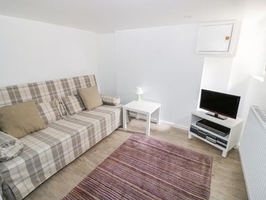 A small living room with a plaid sofa beige cushions a white side table with a lamp a TV on a white stand and a striped rug at Victoria Cottage in Chipping Norton