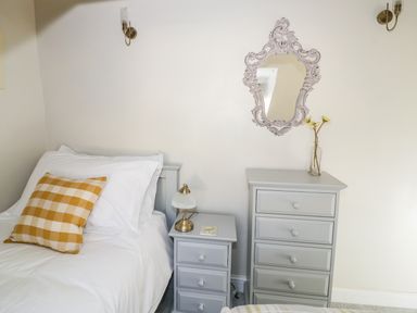 A bedroom with a bed with white linens and a yellow check pillow next to a gray bedside table with a lamp and a coaster and a matching gray tall dresser with a glass vase with flowers and a decorative mirror on the wall at Victoria Cottage in Chipping Norton