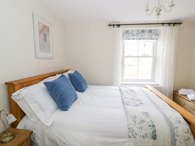 A bedroom with a wooden bed frame blue pillows floral bedding window with patterned roman blind and white curtains at Victoria Cottage in Chipping Norton