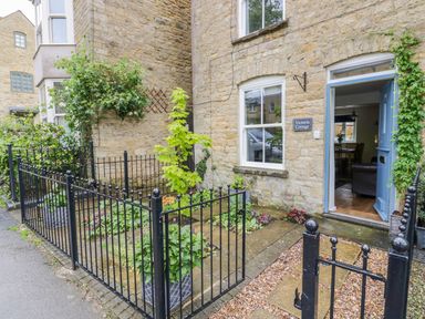Stone exterior of a cottage with a blue door open to a room and a small garden with black iron fencing at Victoria Cottage in Chipping Norton