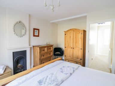 A bedroom with a wooden bed with patterned bedding a wooden dresser wooden wardrobe black chair and a fireplace at Victoria Cottage in Chipping Norton