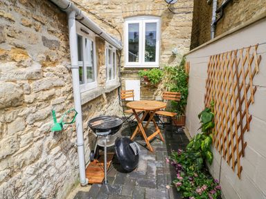 A small outdoor patio with a wooden table and chairs a barbecue and plants at Victoria Cottage in Chipping Norton