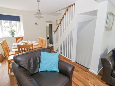 A living room with a dark leather chair and blue cushion next to a wooden dining table with six chairs and tableware at Victoria Cottage in Chipping Norton