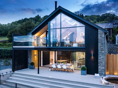 A modern two-story house with large glass windows a dining table on the patio and a hot tub outside at Gatesgarth in Coniston
