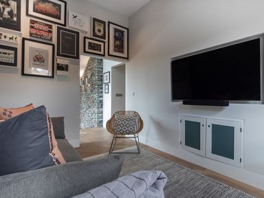 A living room with a gray sofa and pillows a wicker chair a wall-mounted television and framed pictures above a doorway at Gatesgarth in Coniston