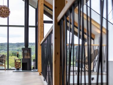 A modern living space with a wood burning stove next to large windows overlooking greenery and a black metal railing with wooden handrail at Gatesgarth in Coniston
