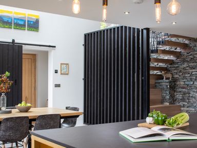 A kitchen and dining area with a wooden table and chairs a book and vegetables on a countertop wooden stairs with stone wall and black slatted divider in Gatesgarth Coniston