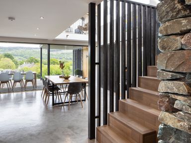 A dining area with wooden tables and chairs next to large glass windows and wooden stairs in a modern home with stone wall at Gatesgarth in Coniston