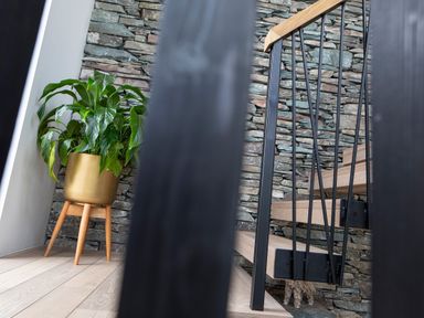 A corner with a green plant in a gold pot and wooden stand next to a wooden staircase with black metal railings and a stone wall at Gatesgarth in Coniston