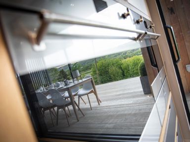 A dining table with chairs on a wooden deck outside a house with glass railing and green trees in the background at Gatesgarth in Coniston
