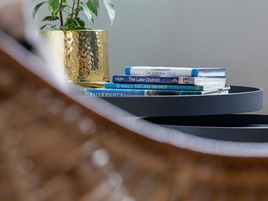 A stack of books and a golden plant pot on a round black table partially obscured by a woven chair at Gatesgarth in Coniston