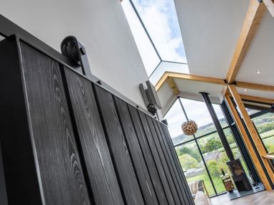 A room with a large window wall showing a green landscape a wood burning stove wooden beams and a black sliding wooden door at Gatesgarth in Coniston