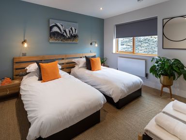 A bedroom with two single beds with white bedding and orange pillows a blue accent wall and a window with a gray blind at Gatesgarth in Coniston