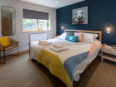 A bedroom with a bed with colorful bedding and pillows a wicker chair with a yellow cushion a window with a blind and a wall mirror at Gatesgarth in Coniston