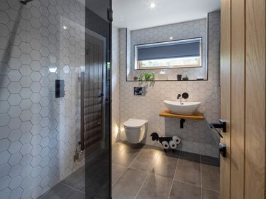 A bathroom with hexagonal wall tiles a floating toilet a sink on a wooden shelf a window with a blind and a glass shower door at Gatesgarth in Coniston