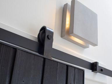A close up of a black sliding door rail with a metal wheel and a square wall light on a white wall at Gatesgarth in Coniston