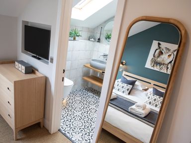 A wooden dresser with a television mounted above next to a bathroom with patterned floor tiles and a wall mirror reflecting a bed with pillows and an owl painting at Gatesgarth in Coniston